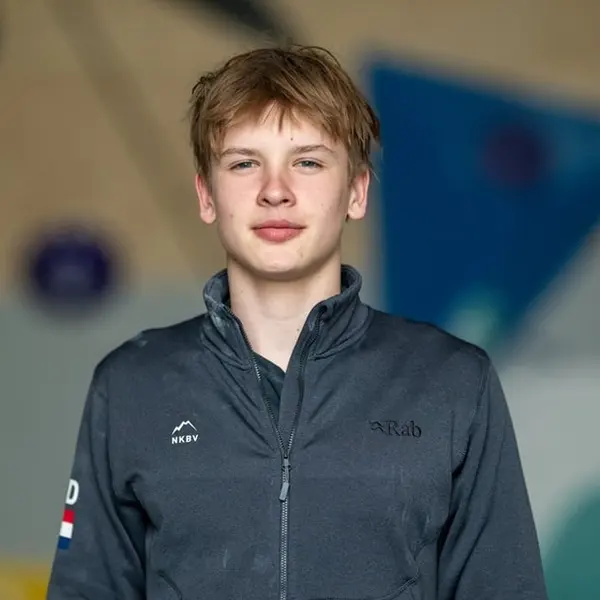 Een tienerjongen met lichtbruin haar draagt een donker jack met ritssluiting en staat binnenshuis, met wazige kleurrijke vormen op de achtergrond.